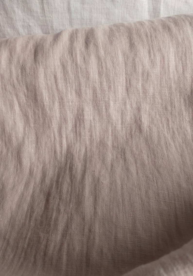 Pode incluir: Um close-up de um tecido de linho marrom claro com um padr&atilde;o tecido texturizado. O tecido est&aacute; dobrado e drapeado, mostrando as rugas e dobras naturais do material.