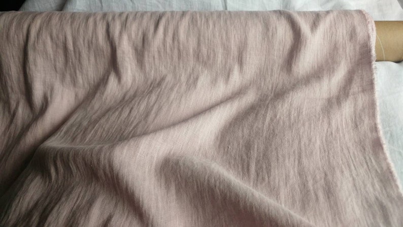 Pode incluir: Um close-up de um tecido macio de cor rosa claro com uma textura sutil. O tecido est&aacute; dobrado e drapeado, mostrando seu caimento e textura.