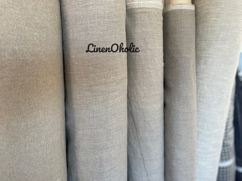 Op de afbeelding: Close-up van verschillende rollen natuurlijke linnen stof. De stof is licht beige van kleur en heeft een licht getextureerd oppervlak. De tekst "LinenOholic" is op een van de rollen gedrukt.