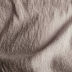 Pode incluir: Um close-up de um tecido de linho macio, de cor castanho claro, com uma textura subtil. O tecido est&aacute; dobrado e drapeado, mostrando o seu caimento natural e as suas rugas.