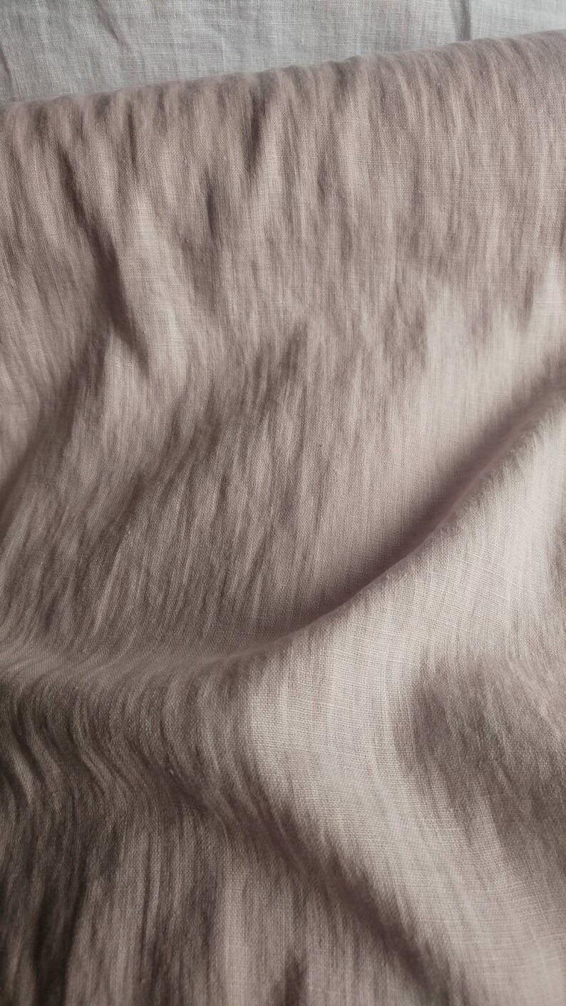 Pode incluir: Um close-up de um tecido de linho macio, de cor castanho claro, com uma textura subtil. O tecido est&aacute; dobrado e drapeado, mostrando o seu caimento natural e as suas rugas.