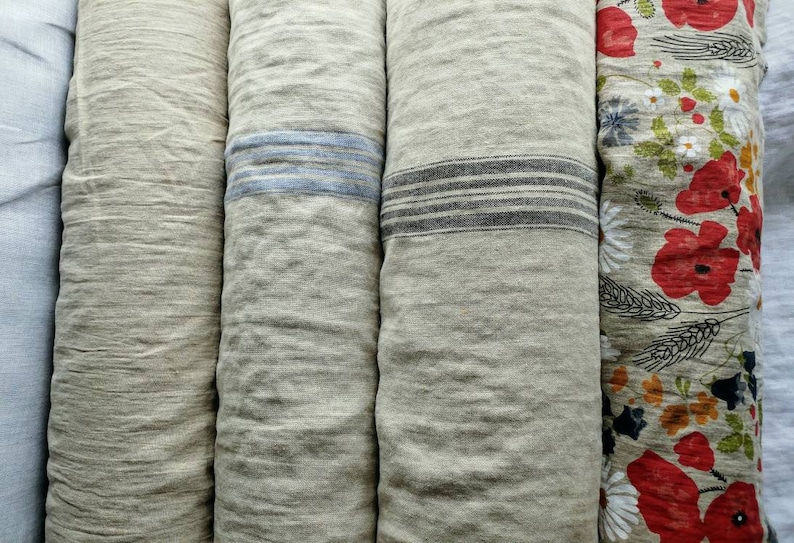 Op de afbeelding: Vier rollen stof, twee met strepen en twee met patronen. De stof is van natuurlijke linnen kleur. E&eacute;n rol heeft blauwe strepen, &eacute;&eacute;n heeft zwarte strepen, &eacute;&eacute;n heeft een bloemenpatroon met rode klaprozen en &eacute;&eacute;n heeft een effen linnen textuur.