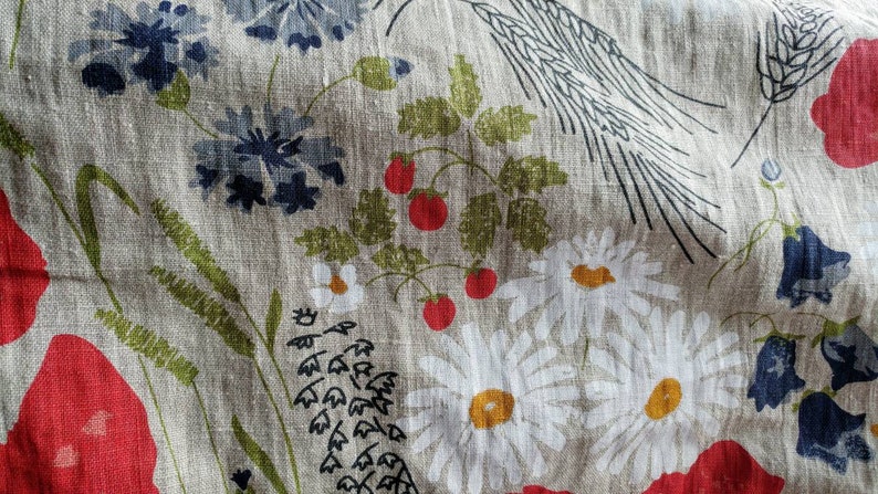 May include: A beige linen fabric with a floral pattern. The pattern includes red poppies, white daisies, bluebells, and green leaves. The fabric has a rustic and vintage feel.