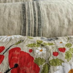 May include: A stack of four linen pillows with different patterns. The top pillow has a floral design with red poppies, white daisies, and green leaves. The second pillow has black and white stripes. The third pillow has a light blue and white pattern. The bottom pillow is a solid beige color.