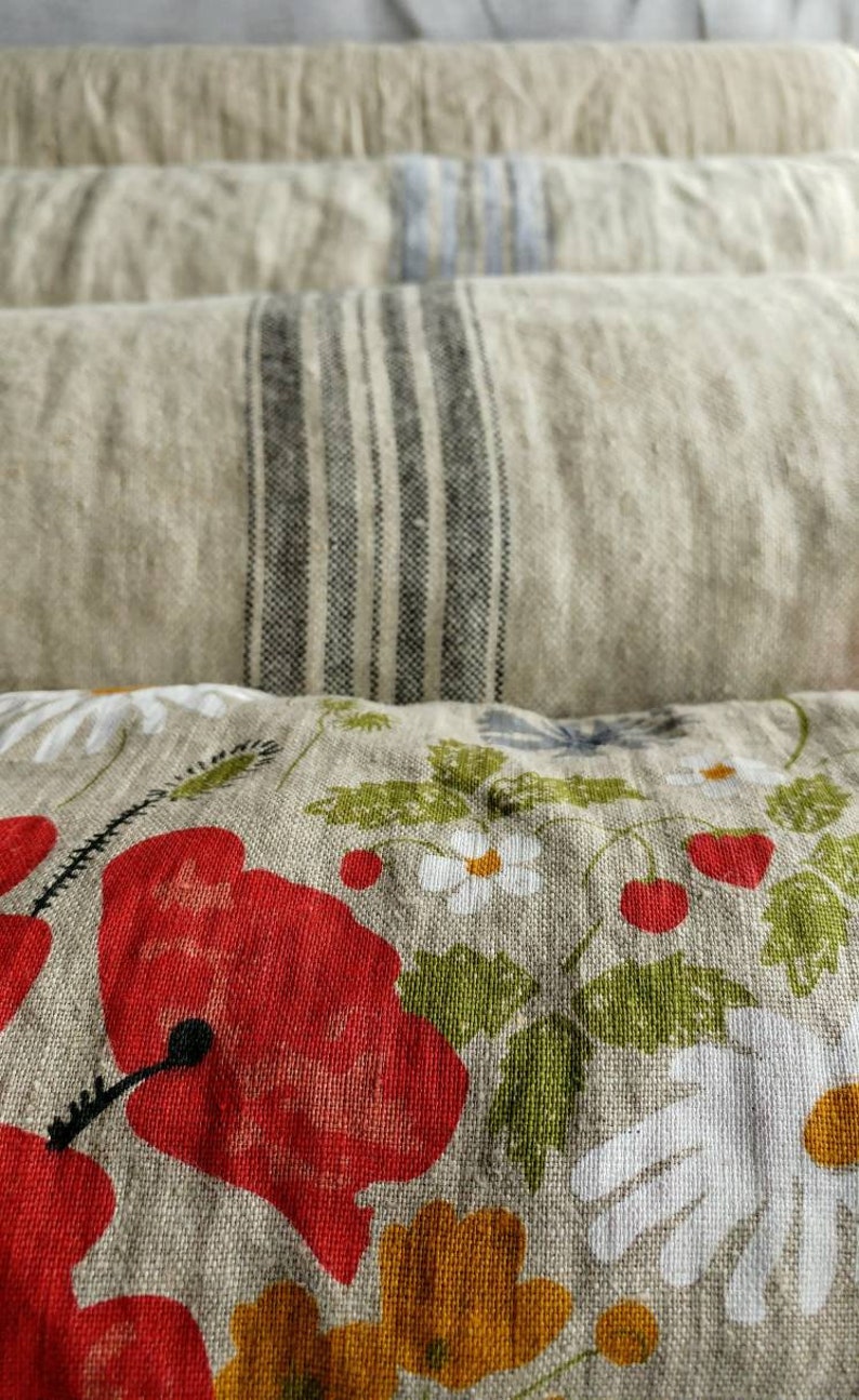 May include: A stack of four linen pillows with different patterns. The top pillow has a floral design with red poppies, white daisies, and green leaves. The second pillow has black and white stripes. The third pillow has a light blue and white pattern. The bottom pillow is a solid beige color.