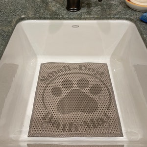 May include: A gray rectangular sink mat with a paw print design and the words "SHOWER DOG" and "PAWESOME" in a white font. The mat is inside a white sink.