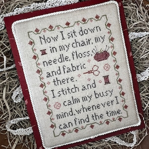 May include: A cross-stitch sampler with a white background and red, green, and brown embroidery. The sampler features a quote about stitching and calming the mind: "Now I sit down in my chair, my needle, floss, and fabric there. I stitch and calm my busy mind, whenever I can find the time."