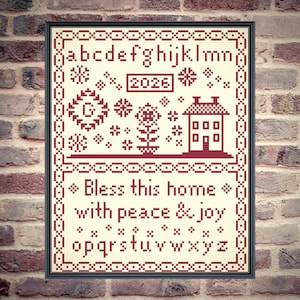 May include: Framed cross-stitch sampler with a cream background and burgundy stitching. The design includes the alphabet, the year 2026, floral and geometric motifs, a house, and the phrase "Bless this home with peace & joy."