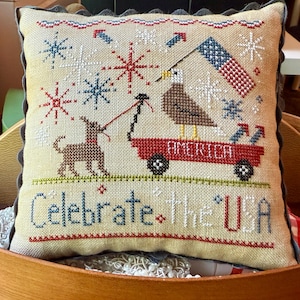 May include: Cross-stitched pillow with a patriotic theme. Features an eagle holding a flag, a dog, and a red wagon with the word "AMERICA". The text "Celebrate The USA" is stitched across the bottom, with snowflake and firework accents.