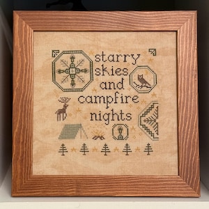 以下が含まれることがあります： ベージュの布地に「starry skies and campfire nights」という文字が刺繍された額入りクロスステッチ。コンパス、フクロウ、シカ、テント、キャンプファイヤー、ランタン、松の木のモチーフがダークグリーンとゴールドの糸で縫い込まれています。