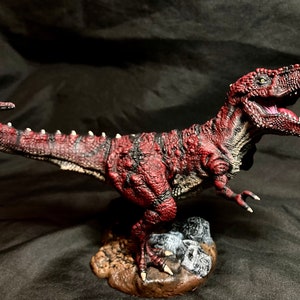 Puede incluir: Una figurita de dinosaurio roja y negra con una superficie texturizada y una boca abierta de par en par. El dinosaurio est&aacute; de pie sobre una peque&ntilde;a base con rocas.