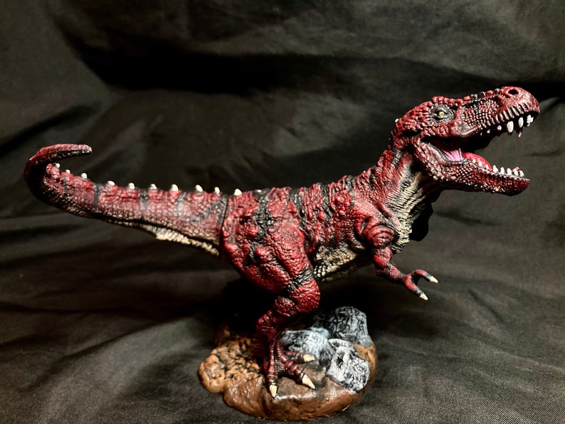 Puede incluir: Una figurita de dinosaurio roja y negra con una superficie texturizada y una boca abierta de par en par. El dinosaurio est&aacute; de pie sobre una peque&ntilde;a base con rocas.