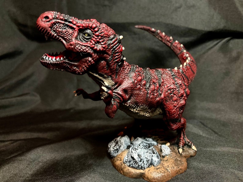 Puede incluir: Una figurita de dinosaurio roja y negra con una superficie texturizada. El dinosaurio est&aacute; en posici&oacute;n de pie con la boca abierta y la cola enroscada alrededor de su cuerpo. La figurita est&aacute; de pie sobre una base con rocas.