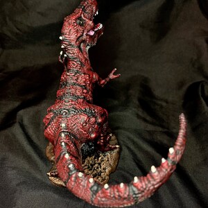 Puede incluir: Una figura de dinosaurio roja y negra con una superficie texturizada y detalles blancos. El dinosaurio est&aacute; en posici&oacute;n de rugido con la cola enroscada alrededor de su cuerpo.