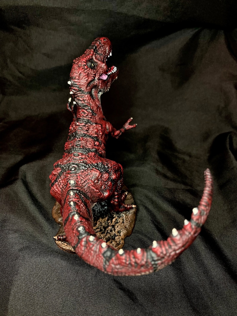 Puede incluir: Una figura de dinosaurio roja y negra con una superficie texturizada y detalles blancos. El dinosaurio est&aacute; en posici&oacute;n de rugido con la cola enroscada alrededor de su cuerpo.