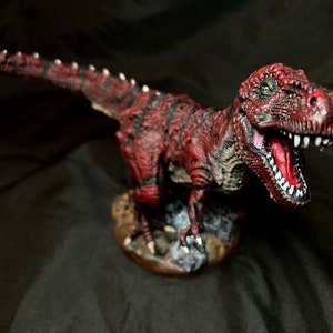 Puede incluir: Una figurita de dinosaurio pintada de rojo y negro con la boca muy abierta, mostrando dientes afilados. El dinosaurio est&aacute; de pie sobre una base marr&oacute;n con textura rocosa.