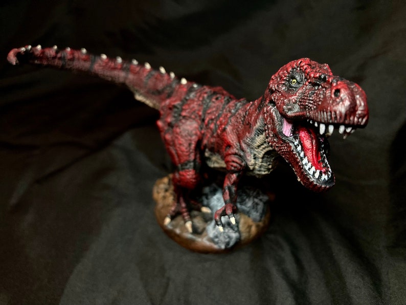 Puede incluir: Una figurita de dinosaurio pintada de rojo y negro con la boca muy abierta, mostrando dientes afilados. El dinosaurio est&aacute; de pie sobre una base marr&oacute;n con textura rocosa.