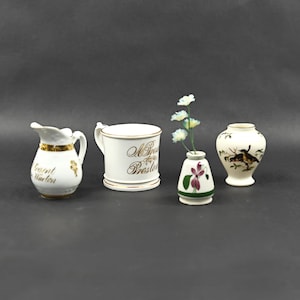 May include: Four white ceramic decorative pieces. A small pitcher with gold trim and text, a mug with gold text, a small vase with flowers, and a vase with a bird design. All are on a gray background.