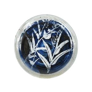 Puede incluir: Plato redondo de cerámica con diseño azul y blanco. El plato presenta un centro azul oscuro con un patrón de hojas blancas. El borde es de color blanquecino moteado. El plato es una pieza decorativa.