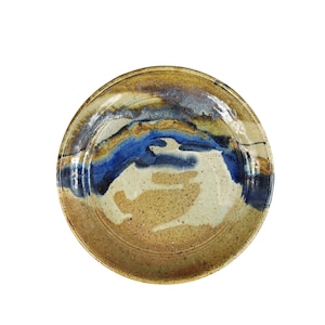 Puede incluir: Un plato de cerámica hecho a mano con un diseño único. El plato presenta una mezcla de colores, incluyendo azul, beige y marrón. El borde tiene un tono marrón más oscuro, y el centro muestra un patrón azul y beige.