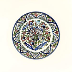 May include: A round, hand-painted ceramic plate with a vibrant floral design. The plate features a peacock in the center, surrounded by red, blue, and green flowers and foliage. The rim is a deep blue, and the plate is set against a white background.