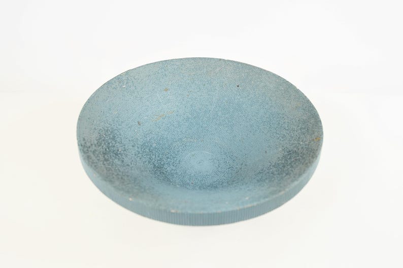 May include: A round, light blue bowl with a textured surface. The bowl has a slightly concave interior and a ridged edge. The color is a muted, dusty blue, and the bowl appears to be made of a solid material.
