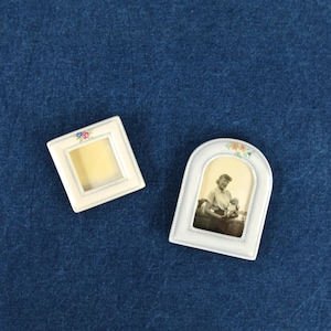 May include: Two white ceramic picture frames with floral accents. One frame is square and the other is arched. The arched frame has a black and white photo of a woman holding a child.