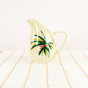 May include: A white ceramic pitcher with a green and yellow floral design. The pitcher has a teardrop shape and a handle.