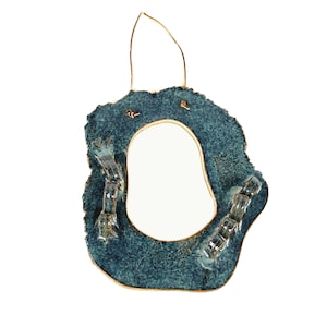May include: A teal ceramic mirror with an irregular shape. It features a gold-coloured trim and a gold wire for hanging. The design includes textured details and small, rectangular embellishments. The mirror's dimensions are approximately 20 cm.