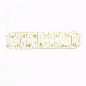 May include: A white ceramic zodiac sign tray with gold zodiac symbols and names. The tray is rectangular with rounded edges and has a hole in the center.