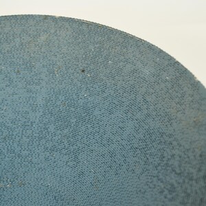 May include: Close-up of a portion of a light blue, circular object with a textured surface. The surface is covered in small, dark dots, creating a pattern. The object is set against a white background.