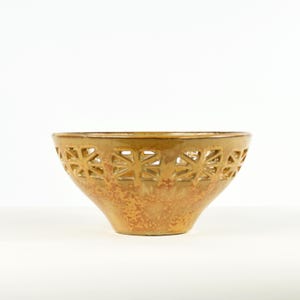 May include: A decorative, ceramic bowl with a warm, earthy color palette. The bowl features a unique design with cut-out geometric shapes around the rim. The bowl's exterior has a textured, speckled appearance, adding to its rustic charm. The bowl is ideal for displaying fruit or as a decorative piece.