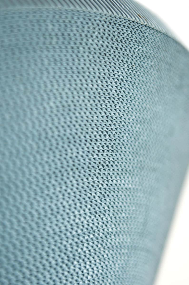 May include: Close-up of a light blue, textured surface with a repeating geometric pattern. The pattern appears to be a series of small, raised triangles, creating a subtle, three-dimensional effect. The surface curves slightly, suggesting a cylindrical or rounded shape.