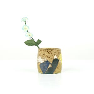 May include: A speckled ceramic vase with a mountain design in blue, white, yellow, and green. The vase holds a sprig of light blue flowers. The vase is cylindrical and has a rustic, handmade appearance.