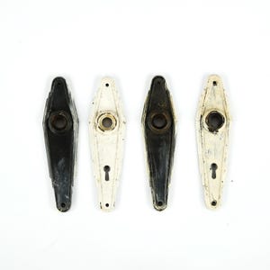 May include: Four vintage door handle backplates in black and off-white. Each backplate has a keyhole and a central circular opening. The handles have a geometric, Art Deco design and show signs of wear.