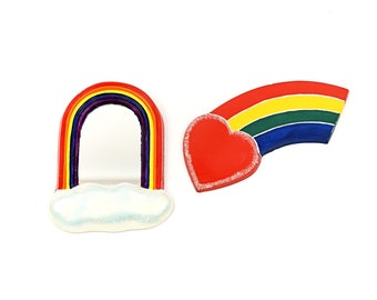 May include: Two rainbow-colored heart-shaped magnets. One rainbow magnet is shaped like an arch with a white cloud at the bottom. The other rainbow magnet is shaped like a half-circle with a red heart at the top.