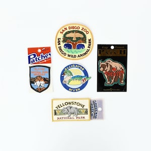 Può includere: Una collezione di cinque toppe da stirare che rappresentano diversi parchi nazionali e animali selvatici. Le toppe sono colorate e presentano immagini di un orso, un'anatra, un bisonte, una catena montuosa e un albero con animali. Le toppe sono etichettate con i nomi dei parchi e degli animali selvatici che rappresentano, tra cui "Glacier National Park Montana", "Yellowstone National Park", "Alligator River", "Rockies" e "San Diego Zoo Wild Animal Park".