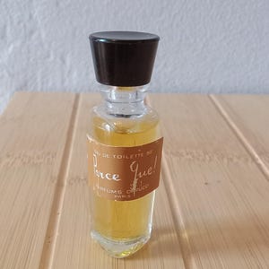 May include: A small, clear glass perfume bottle filled with amber-colored liquid, featuring a black cap and a tan label with white text that reads "Parce Que! Eau de Toilette 80 Parfums O. Pucci Paris" in a script and sans-serif font.