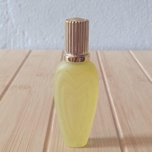 May include: A small, frosted yellow glass perfume bottle with a teardrop shape and embossed heart patterns on the surface. The bottle features a metallic gold cylindrical cap with vertical ridges and a smooth, rounded top finish.