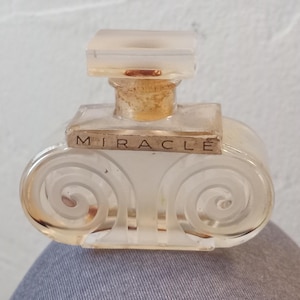 May include: A clear glass perfume bottle with a square top and a decorative, swirled design on the sides. The word "MIRACLE" is printed on a small label. The bottle has a light brown cork and a clear, rectangular stopper.