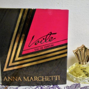 May include: A vintage perfume bottle and box. The box is black with gold accents and a pink top, featuring the text "Voile Eau de Parfum" and "Anna Marchetti." The perfume bottle is a clear, geometric shape with a gold-colored cap.