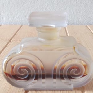 May include: A clear, antique perfume bottle with a rectangular stopper. The bottle has a rounded shape with etched spiral designs on the front. The liquid inside is amber-colored. The bottle is displayed on a light-colored wooden surface.