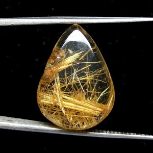May include: A teardrop-shaped gemstone with golden, needle-like inclusions suspended within a clear, amber-colored body. The gem is held by silver tweezers against a black background, highlighting its clarity and internal structure.