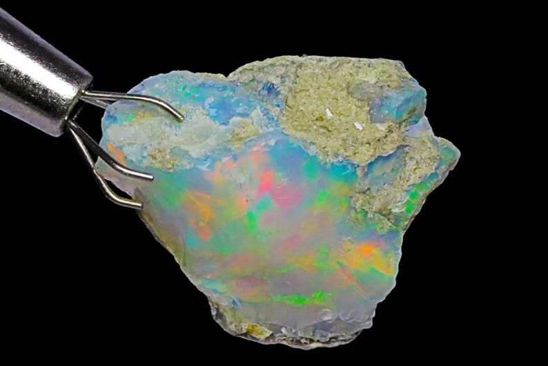 May include: A rough, natural opal gemstone with a vibrant iridescent display of green, blue, orange, and red colors. The opal is held by a metal tool.