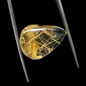 May include: A clear, teardrop-shaped gemstone with golden, needle-like inclusions, held by silver tweezers against a black background. The gemstone has a polished surface and a small notch at the top.