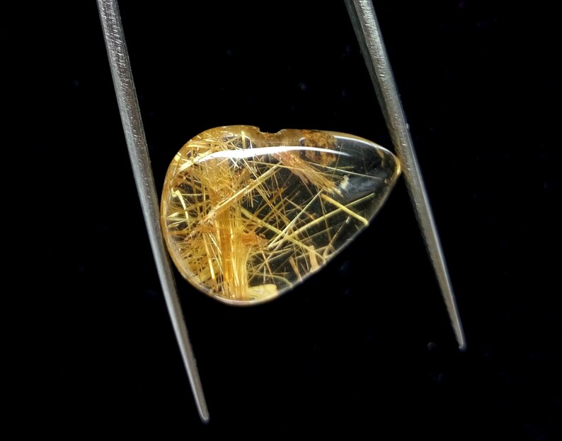 May include: A clear, teardrop-shaped gemstone with golden, needle-like inclusions, held by silver tweezers against a black background. The gemstone has a polished surface and a small notch at the top.
