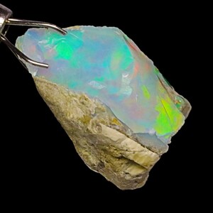 May include: A rough, natural opal gemstone with a light blue, green, and orange iridescent sheen. The opal is held by a metal tool.