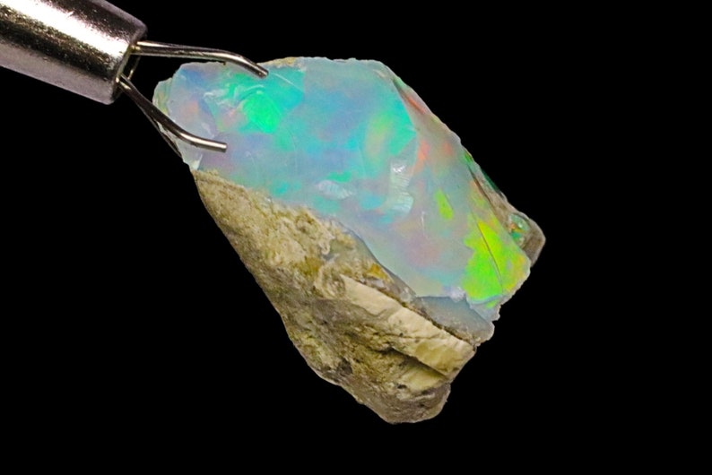 May include: A rough, natural opal gemstone with a light blue, green, and orange iridescent sheen. The opal is held by a metal tool.