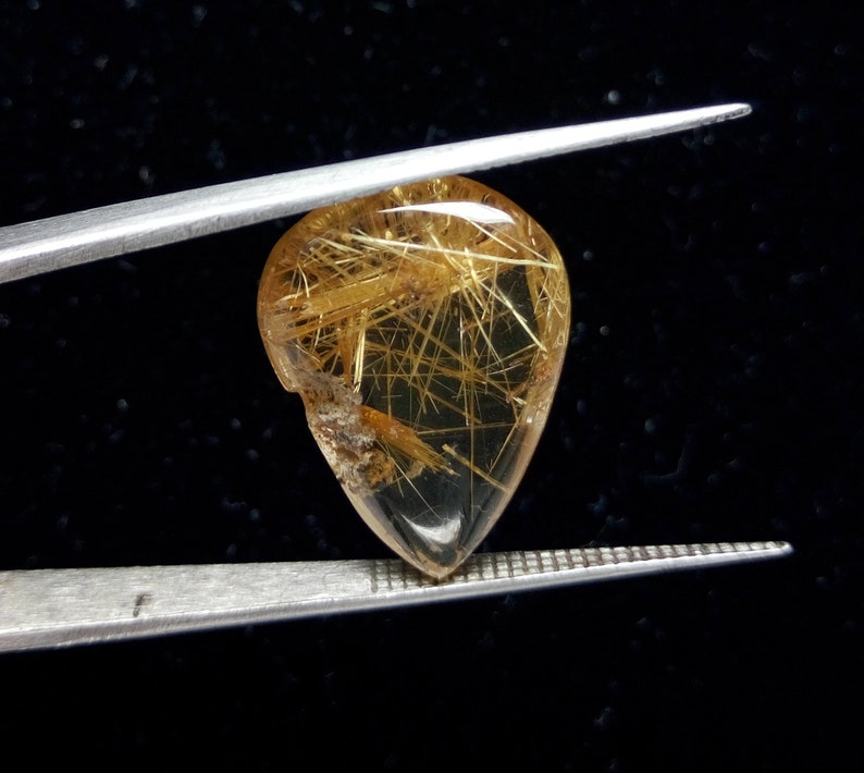 May include: A teardrop-shaped gemstone with golden, needle-like inclusions, held by silver tweezers against a black background. The gem is translucent with a polished surface, showcasing the internal structures.