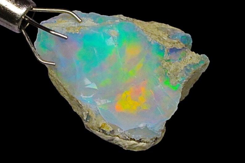 May include: A rough, natural opal gemstone with a milky white base and iridescent flashes of green, blue, orange, and yellow.